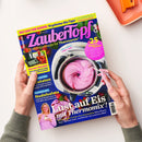 Zwei Hände halten eine schräg liegende Zeitschrift mit dunkelblauem Hintergrund und Erdbeeren vor weißer Fläche. Auf dem Titel stehen „Zaubertopf“, „Lieblingsrezepte für Thermomix®“, „DAS SAGT DIR KEINER: 10 geheime Mix-Tipps“ und „+25 Express-Rezepte“; abgebildet sind ein Thermomix-Gerät mit rosa Speise, Rezeptfotos und eine Frau.