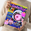Weibliche Person im beigefarbenen Strickpullover hält ein schräg fotografiertes Magazin mit blau-gelbem Cover. Darauf: pinke Eiscreme im Thermomix®-Mixtopf, Erdbeeren und ein Mixer; lesbare Texte sind „DAS SAGT DIR KEINER: 10 geheime Mix-Tipps“, „Lieblingsrezepte für Thermomix®“, „5 Zutaten“, „Nudelsalate“ und „Lust auf Eis mit Thermomix!“.