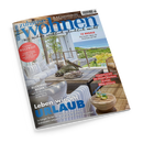 Schräg fotografierte Wohnzeitschrift „Zuhause wohnen“ mit glänzendem Cover: helle Terrasse mit weißem Sofa, Holztisch, Korbsessel, Pflanzen, Getränken und Meerblick. Sichtbare Texte: „Wie Wohnen wirklich glücklich macht“, „Leben wie im URLAUB“, „Genuss WELTWEIT“, „Blickfang Bilder an der Wand“, „Kaffee & Eis“ und „Die neuesten Maschinen & Trends“.
