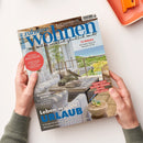 Schräg auf einer weißen Fläche gehaltene Wohnzeitschrift „neue zuhause WOHNEN“ mit blauem Titel und dem Text „Wie Wohnen wirklich glücklich macht“. Das Cover zeigt eine helle Terrasse mit Meerblick sowie die Aufschriften „12 REGale“, „Genuss WELTWEIT“, „BLICKFANG BILDER“, „Leben wie im URLAUB“ und „KAFFEE & EIS“; zwei Hände mit grauen Strickärmeln halten das Heft.